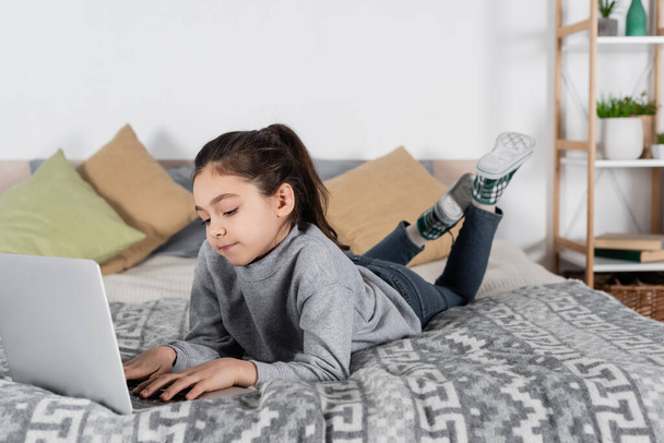 full length view of preteen girl lying on bed and typing on laptop - 写真・画像