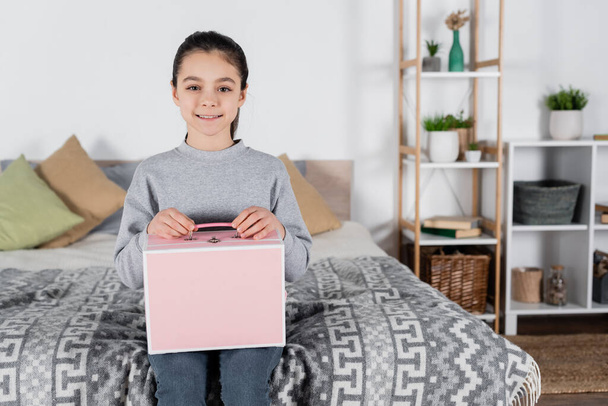 cheerful girl looking at camera while sitting on bed with pink box - Fotó, kép