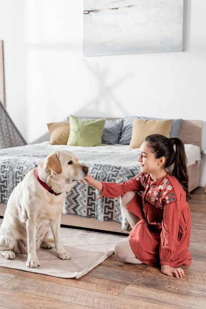 smiling girl stroking labrador dog while sitting on floor in bedroom - Photo, Image