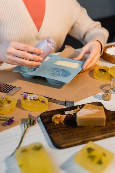 Vista recortada de artesana sosteniendo tarro con jabón líquido cerca de molde de silicona y flores secas en la mesa  - Foto, Imagen