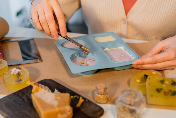 Vista recortada de la artesana vertiendo azafrán sobre jabón en molde de silicona sobre papel artesanal sobre mesa - Foto, Imagen