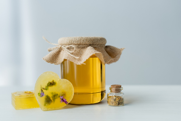 Frasco com mel perto de saboneteiras artesanais e ervas na mesa em fundo cinza  - Foto, Imagem
