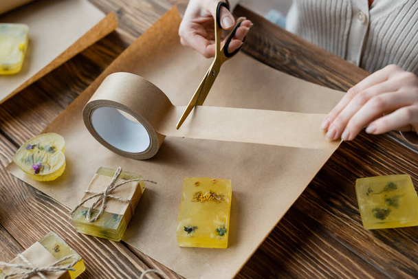 Oříznutý pohled na řemeslnici stříhající lepicí pásku v blízkosti ručně vyrobeného mýdla na řemeslném papíru  - Fotografie, Obrázek