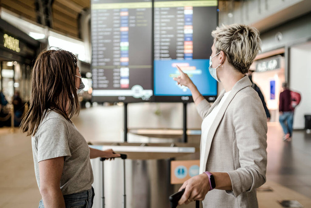 Two travelling women wearing protective masks discussing by flight information board at the Faro airport - Photo, Image