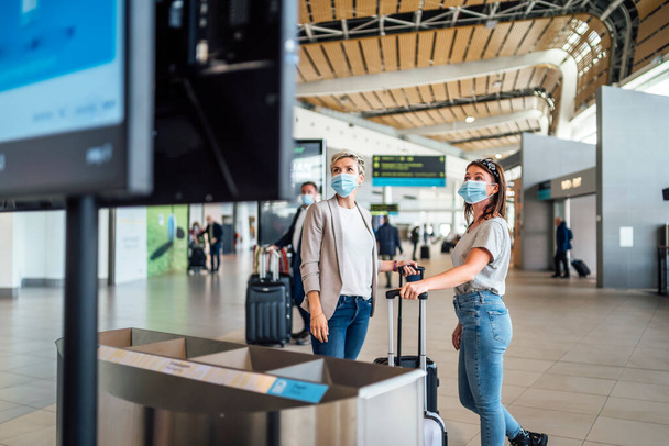 Δύο γυναίκες που ταξιδεύουν φορώντας προστατευτικές μάσκες συζητώντας μέσω του σκάφους πληροφοριών πτήσης στο αεροδρόμιο Faro - Φωτογραφία, εικόνα
