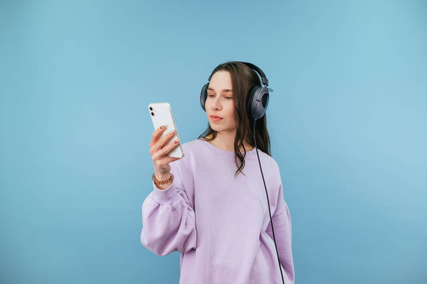 Ελκυστική γυναίκα με ακουστικά και μωβ φούτερ στέκεται σε μπλε φόντο και χρησιμοποιεί ένα smartphone με σοβαρό πρόσωπο. - Φωτογραφία, εικόνα