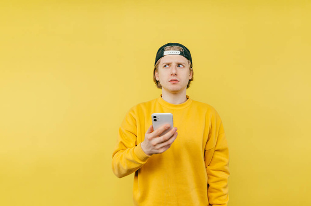 Νεαρός άνδρας με κίτρινη μπλούζα στέκεται με ένα smartphone στα χέρια του σε κίτρινο φόντο και κοιτάζει αλλού με σοβαρό πρόσωπο. - Φωτογραφία, εικόνα