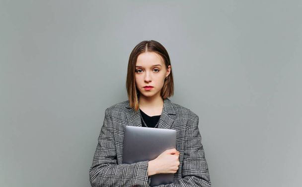 Ελκυστική επιχειρηματίας με έξυπνα casual ρούχα με ένα φορητό υπολογιστή στα χέρια της στέκεται σε ένα γκρι φόντο και κοιτάζει την κάμερα με ένα σοβαρό πρόσωπο. - Φωτογραφία, εικόνα