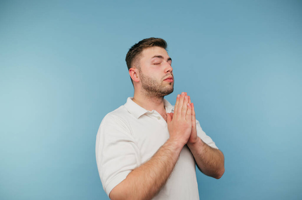 Un hombre con una camiseta blanca reza a Dios con los ojos cerrados y los brazos cruzados sobre un fondo azul. - Foto, Imagen