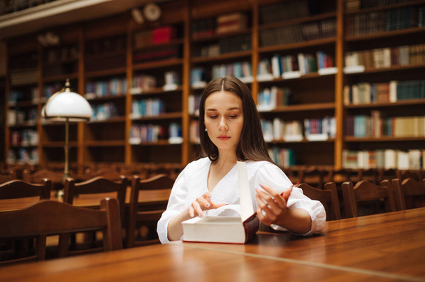 Vonzó női diák könyvet olvas, miközben egy komoly arcú asztalnál ül az egyetemi egyetemi egyetemi könyvtárban.. - Fotó, kép