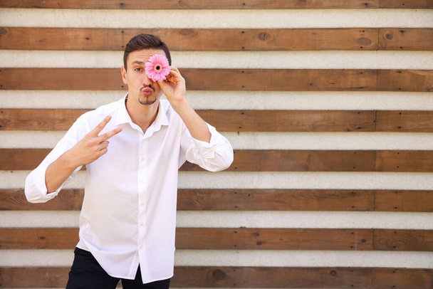 Lustiger kaukasischer Typ in weißem Hemd mit V-Zeichen und küssenden Lippen, der eine rosa Blume anstelle eines Auges auf einem braunen gerippten Hintergrund mit Kopierraum hält - Foto, Bild