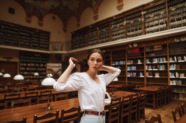 Schöne Studentin in weißer Bluse posiert mit ernstem Gesicht in der alten atmosphärischen Bibliothek vor der Kamera - Foto, Bild