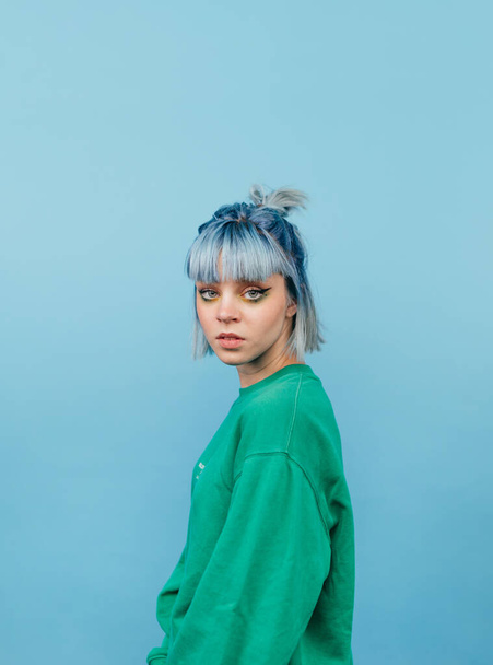 Portrait of a beautiful woman with blue hair posing at camera on a blue background with a serious face in a green sweatshirt. - Photo, Image