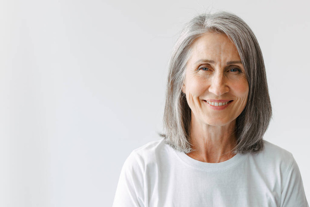 Grey senior woman in t-shirt smiling and looking at camera isolated over white background - Zdjęcie, obraz