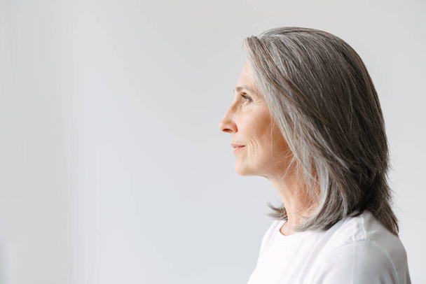 Серая старшая женщина в футболке смотрит в сторону, позируя в профиль изолированы на белом фоне - Фото, изображение