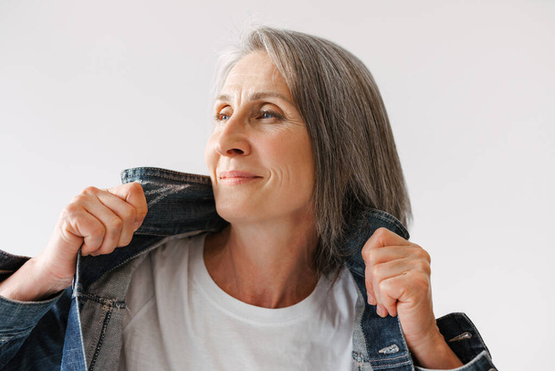 Grey senior woman smiling while posing in denim jacket isolated over white background - 写真・画像