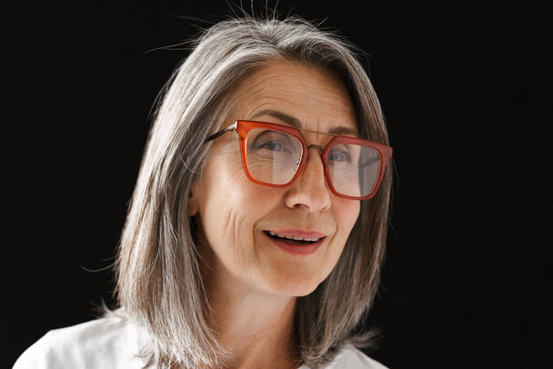Grey senior woman in eyeglasses smiling and looking aside isolated over black background - Zdjęcie, obraz