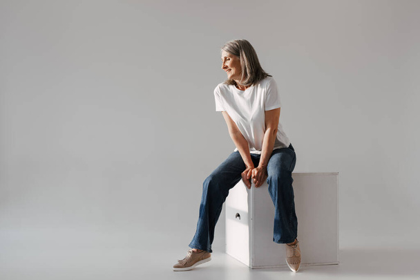 Grey senior woman in jeans smiling while sitting on box isolated over white background - Фото, изображение