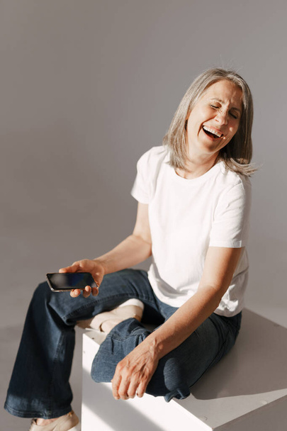 Grey senior woman using mobile phone while sitting on box isolated over white background - Foto, Imagem