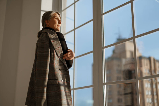 European senior woman with grey hair looking out window indoors - Foto, imagen