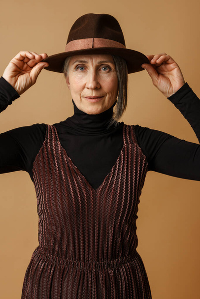 Grey senior woman holding her hat and looking at camera isolated over beige background - Foto, imagen