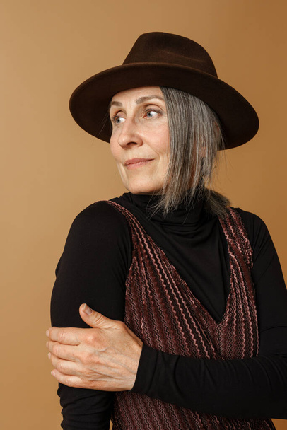 Grey senior woman in hat posing and looking aside isolated over beige background - Foto, immagini