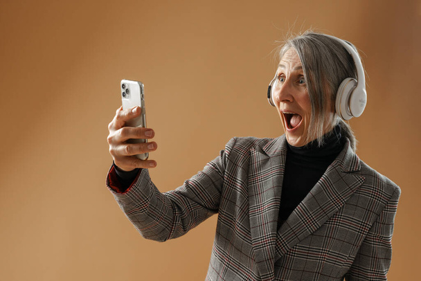 Grey senior woman in jacket using headphones and cellphone isolated over beige background - Foto, Imagen