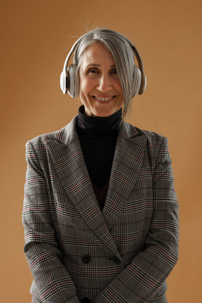 Grey senior woman in headphones smiling and looking at camera isolated over beige background - Foto, Imagen