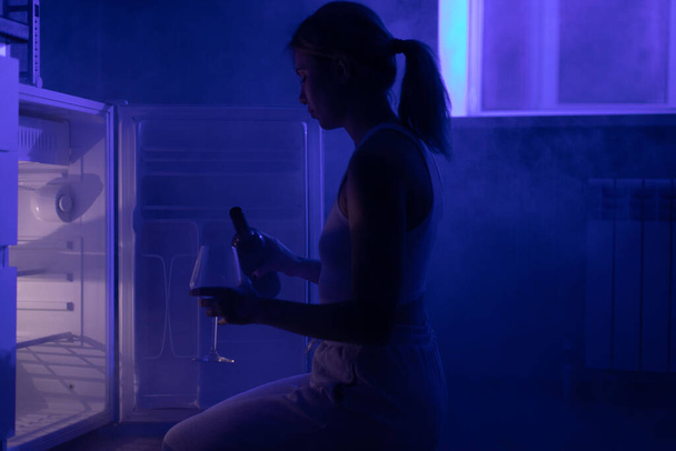 Sad woman drinking wine sitting in front of an open refrigerator in the dark. - Foto, Imagen
