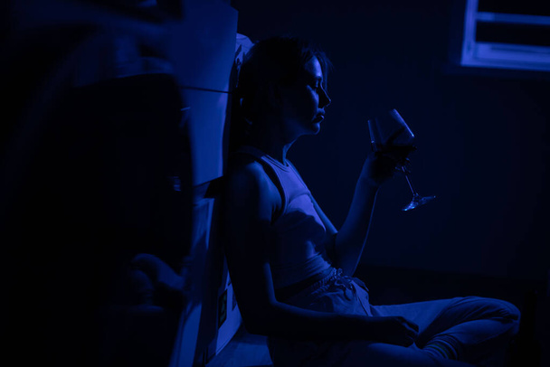 Sad woman drinking wine while sitting near the refrigerator in the dark. - Фото, изображение