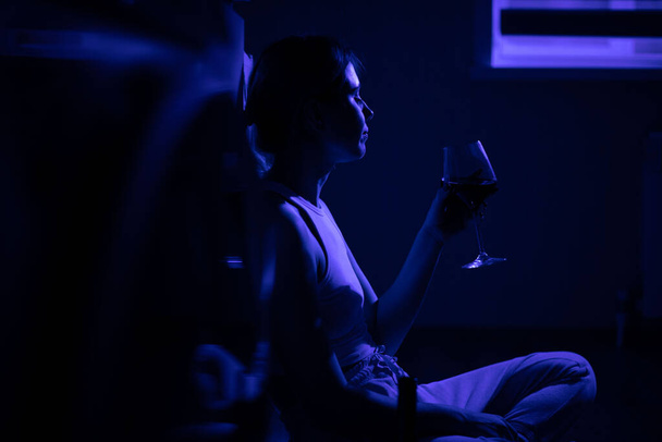 Sad woman drinking wine while sitting near the refrigerator in the dark. - Zdjęcie, obraz