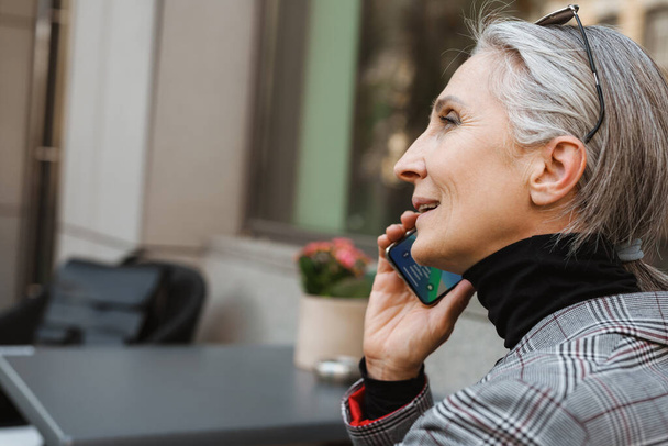 Szürke fehér idős nő beszél mobilon, miközben a szabadban ül egy kávézóban. - Fotó, kép