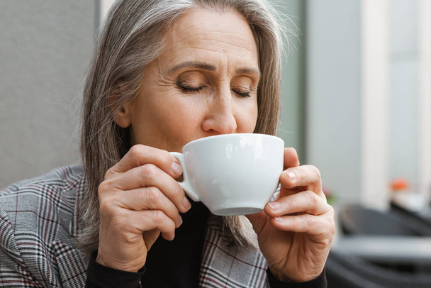Серо-белая пожилая женщина пьет кофе, сидя в кафе на открытом воздухе - Фото, изображение