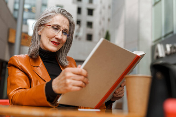 Сіра біла старша жінка читає книгу, сидячи в кафе на відкритому повітрі
 - Фото, зображення