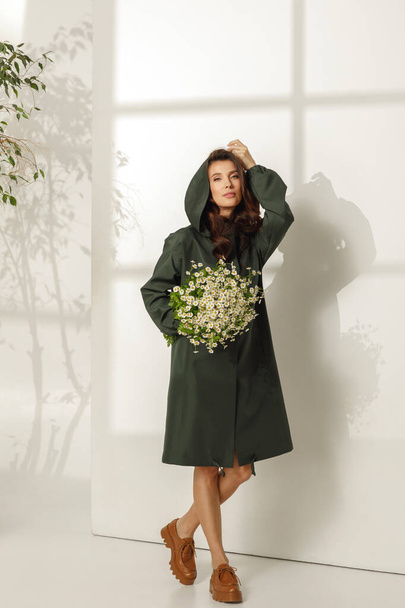 Das weibliche Model trägt ein trendiges Frühlingsoutfit, einen khakifarbenen Regenmantel und hält einen Strauß Gänseblümchen vor weißem Hintergrund. Frühling, Sommer Modekonzept - Foto, Bild