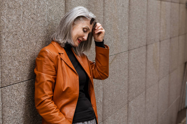 Grey senior woman in leather jacket smiling while leaning on wall outdoors - Photo, Image