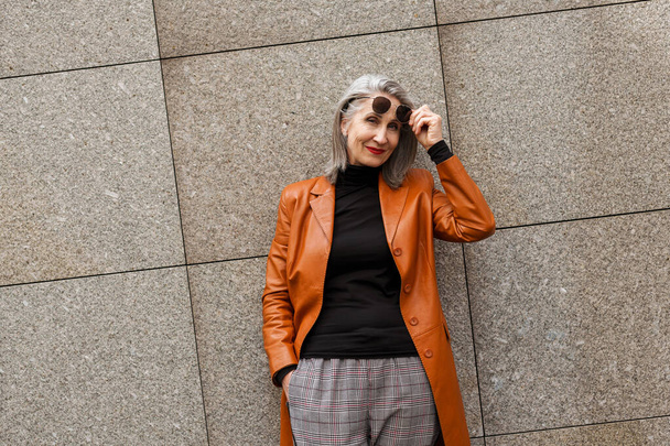 Grey senior woman in leather jacket smiling at camera while leaning on wall outdoors - Fotografie, Obrázek