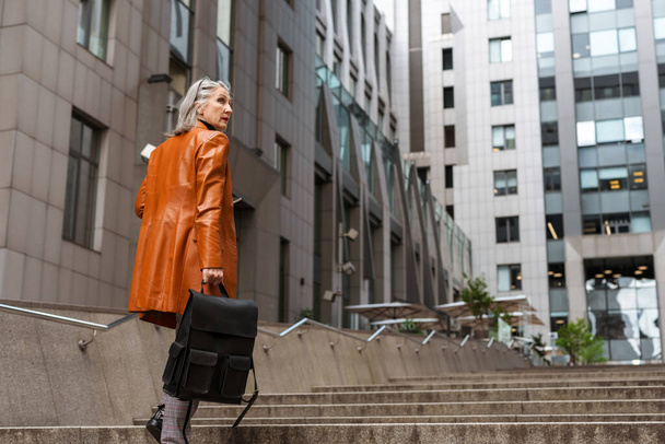 Grey senior woman in leather jacket holding bag while going up stairs outdoors - Foto, afbeelding