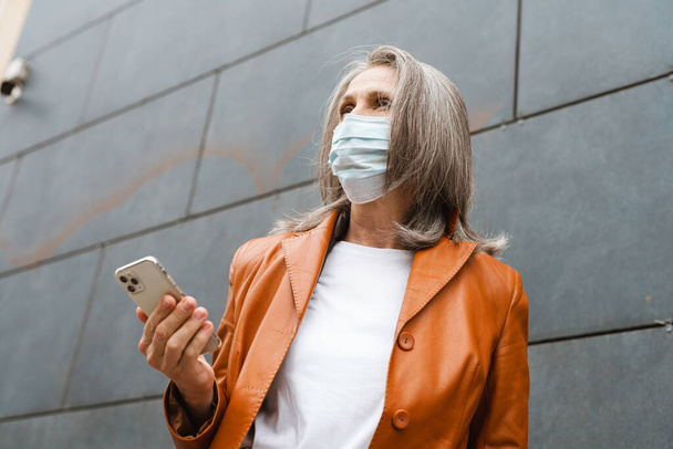 Grey senior woman in face mask using mobile phone outdoors - Photo, Image