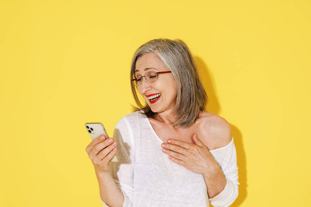 Mature woman in eyeglasses laughing while using mobile phone isolated over yellow wall - Valokuva, kuva