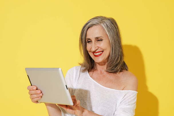 White senior woman with grey hair smiling while using tablet computer isolated over yellow wall - Fotó, kép