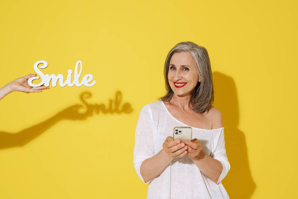 Mature woman using cellphone while posing with decorated white word isolated over yellow wall - Photo, image