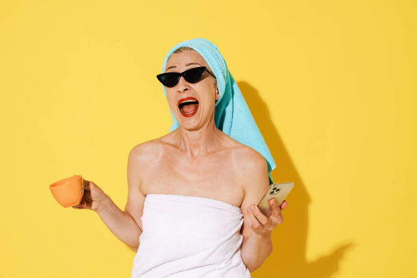 Mature woman in sunglasses using cellphone and drinking coffee isolated over yellow wall - Zdjęcie, obraz