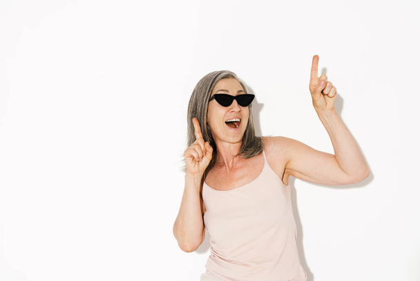 Senior woman wearing sunglasses laughing while pointing fingers upward isolated over white wall - Φωτογραφία, εικόνα