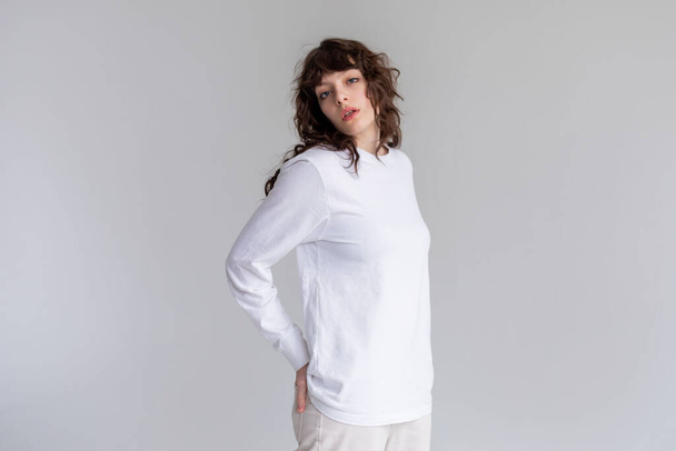 Mujer joven con el pelo rizado en una camiseta blanca de manga larga está de pie sobre un fondo blanco. Mock-up. - Foto, imagen