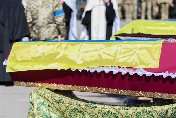 POLTAVA, UKRAINE - MARCH 17, 2022: Red wooden coffins covered with Ukrainian flags with victims of Russian attack on Ukraine during funeral service - Photo, Image