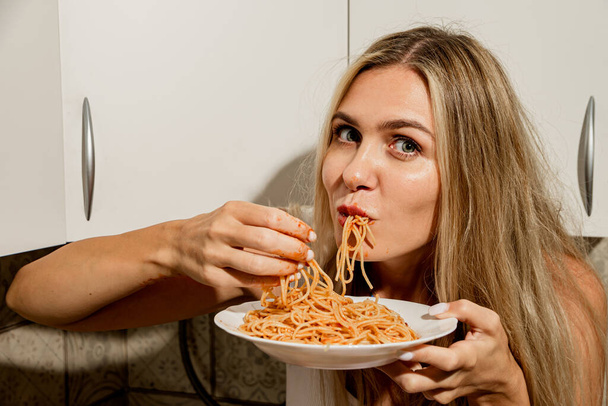 Gedurfd sexy langbenige schoonheid in een wit bodysuit eet spaghetti met tomatenpasta met haar handen. Moderne huisvrouw eet pasta in een moderne Scandinavische keuken, zit op de keukentafel - Foto, afbeelding