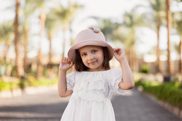 Portrait of a happy little girl in white boho dress and straw hat on the background of palm trees. - Zdjęcie, obraz