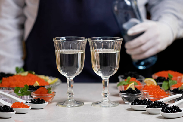 Kelner nalewający szampana lub białe wino z butelki do szklanek przy stole w formie bufetu podczas degustacji przystawek z czerwonym i czarnym kawiorem. Degustacja łososia czerwony kawior i czarny jesiotr beluga solone ikra. Zestaw pysznych przekąsek na bankiecie - Zdjęcie, obraz