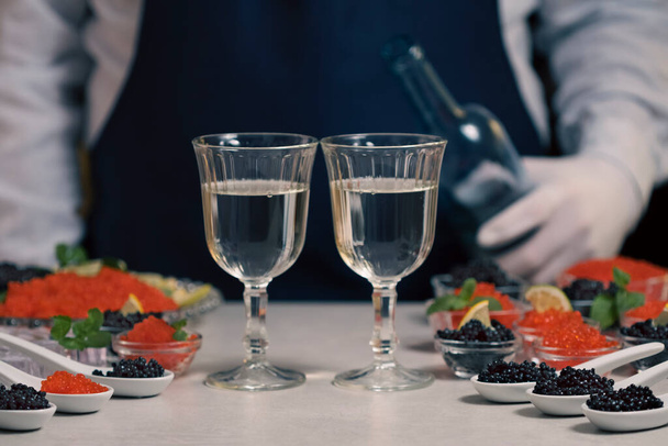 Kelner nalewający szampana lub białe wino z butelki do szklanek przy stole w formie bufetu podczas degustacji przystawek z czerwonym i czarnym kawiorem. Degustacja łososia czerwony kawior i czarny jesiotr beluga solone ikra. Zestaw pysznych przekąsek na bankiecie - Zdjęcie, obraz